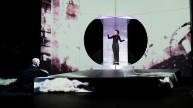 : Eine Wand mit einer sehr hellen Projektion, in der Mitte eine kreisrunde Öffnung, in der eine schwarzgekleidete, singende Frau steht. Vor ihr ein weißes, niedriges Podest. Links vorne sitzt ein glatzköpfiger Mann am Boden und sieht ihr zu, die Arme auf das Podest abgelegt.