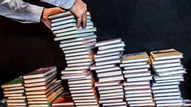 Zwei Hände legen Bücher auf einen bereits hochen Buchstapel.