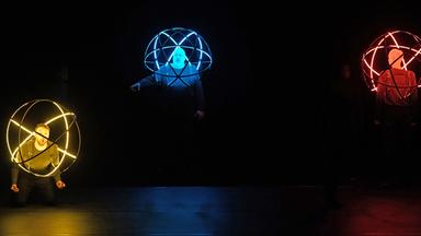 Ein schwarzer Hintergrund. Zu sehen sind drei Solisten, die mittels einer großen Lichtkugel, die sie auf ihrem Oberkörper tragen, in gelbes, blaues und rotes Licht gehüllt werden.