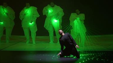 Ein Mann kniet auf der Bühne. Hinter ihm stehen vier Tänzer in grünes Licht gehüllt. Lasterstrahlen sind auf den Mann im Vordergrund gerichtet