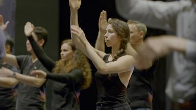 Mehrere schwarzgekleidete Tänzerinnen und Tänzer bei der Probe. Sie halten jeweils den rechten Arm leicht angeinkelt nach oben. Der linke Arm ist jeweil auf Brusthöhe nach vorne gestreckt. 