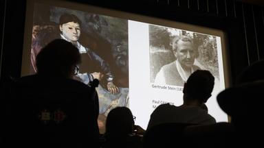 Menschen in einem dunklen Raum betrachten an die Wand projizierte Fotos von einem Porträt-Gemälde und einer historischen Fotografie von einem Mann.