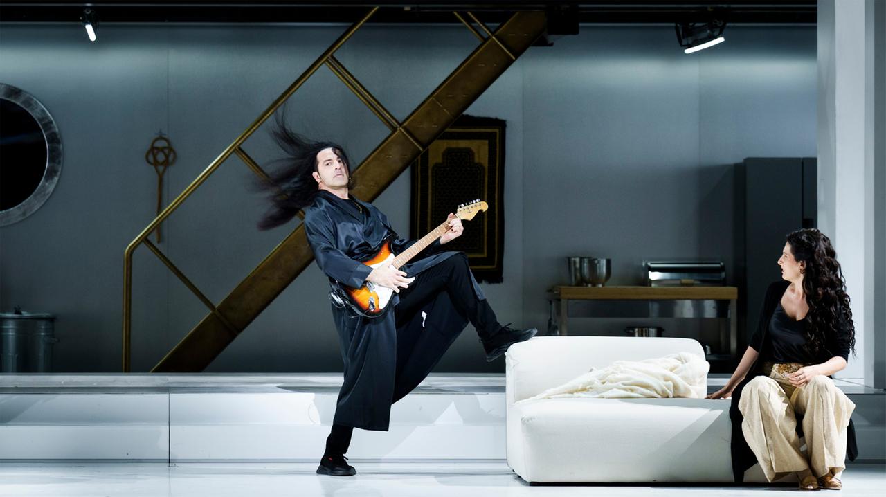 Eine Person mit einer E-Gitarre steht auf einer Theaterbühne vor einer modernen Kulisse mit Treppe und weißem Sofa. Eine weitere Person sitzt auf dem Sofa und blickt zur Bühnenmitte.