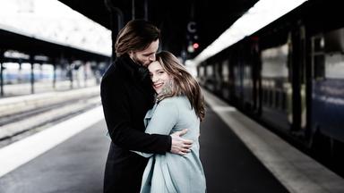 Ein Mann und eine Frau stehen an einem Bahnsteig, umarmen sich und er gibt ihr einen Kuss auf die Stirn.