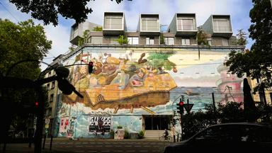 Eine hohe Gebäudefassade ist mit einem farbenfrohen Wandgemälde verziert. Das Mural zeigt Menschen und surreale Figuren in einem riesigen Pappkarton auf hoher See. 