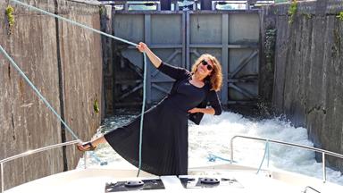 Moderatorin Simin Sadeghi im schwarzen Kleid und mit Sonnenbrille steht auf dem Hausboot  und hebt lachend ein Bein in die Luft