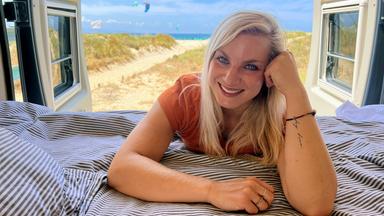 Eine blonde Frau lehnt mit dem Oberkörper auf einer Matratze mit Bettzeug im hinteren Teil eines Campervans. Im Hintergrund ein Strand und das Meer.