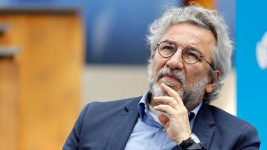 Foto eines Mannes mit weißen Locken, graumeliertem Bart und runder Metall-Brille, der sich nachdenklich mit der Hand ans Kinn fasst.