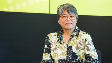 Foto einer asiatisch aussehenden Frau mittleren Alters mit kurzen graumelierten Haaren und Brille, die ein grün, weiß, grau gemustertes Hemd trägt.