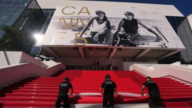 Drei schwarz gekleidete Männer rollen auf den Treppen zum Kino in Cannes einen roten Teppich ab.