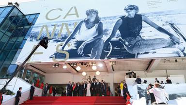 Vor dem Eingang zum Festivalkino in Cannes steht eine Gruppe Menschen. Über ihnen ist das riesige Plakat des Filmfestivals Cannes zu sehen.