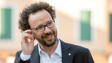 Ein lächelnder Mann mit lockigem, brauen Haar, einer Brille und Anzug.