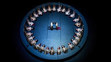 Menschen liegen in einem Kreis auf dem Boden. In der Mitte des Kreises knien sich zwei Menschen gegenüber und berühren sich mit den Köpfen.