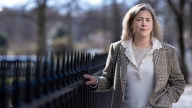 Porträtfoto einer Frau mit schulterlangen Haaren, die einen karierten Anzug trägt. Sie steht an einem schmiedeeisernen Zaun und blickt ernst in die Kamera.