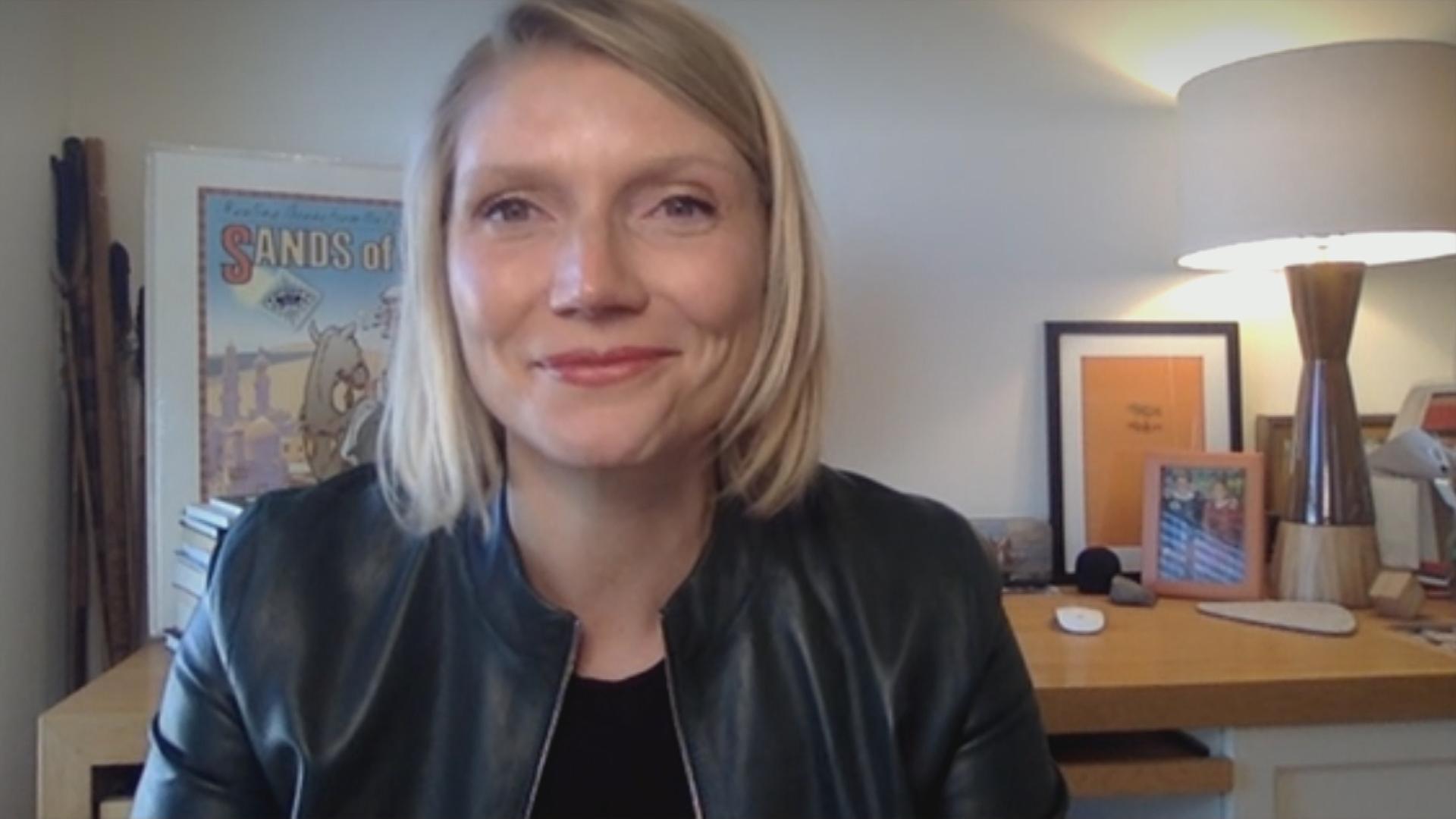 Eine Frau mit blonden Haaren und Lederjacke sitzt in einem Zimmer und lächelt in die Kamera.