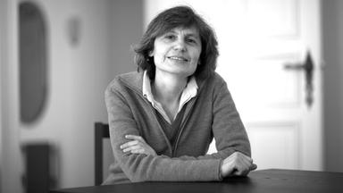 Das schwarz-weiß Foto einer Frau, die an einem Tisch sitzt, die Arme vor sich verschwänkt und in die Kamera lächelt.