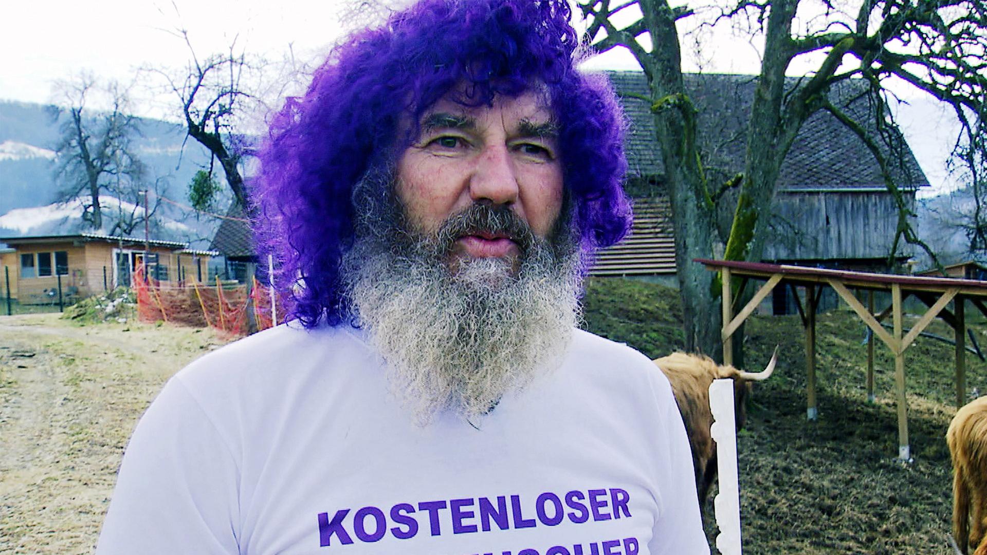 Das Bild zeigt einen Mann mit einem markanten Aussehen. Er hat einen dichten, grauen Bart und trägt eine hellgraue T-Shirt mit dem Aufdruck „KOSTENLOSER KURPFUSCHER“ in lila Buchstaben. Seine Haare sind auffällig lila gefärbt und lockig. Der Mann steht in einem Außenbereich, hinter ihm sind mehrere Hochlandrinder zu sehen, die dichtes, hellbraunes Fell haben. Die Umgebung ist ländlich, mit einem leicht bewaldeten Hintergrund und einem Holzgebäude, das in der Ferne sichtbar ist. Um den Bereich sind Absperrungen zu erkennen, und der Boden ist überwiegend braun mit kurzer Vegetation. Der Himmel ist bewölkt, was auf schlechtes Wetter hindeutet. Die gesamte Szene vermittelt eine entspannte, jedoch ungewöhnliche Atmosphäre, die mit der Thematik der Energetik in Verbindung steht, die im Text erwähnt wird.