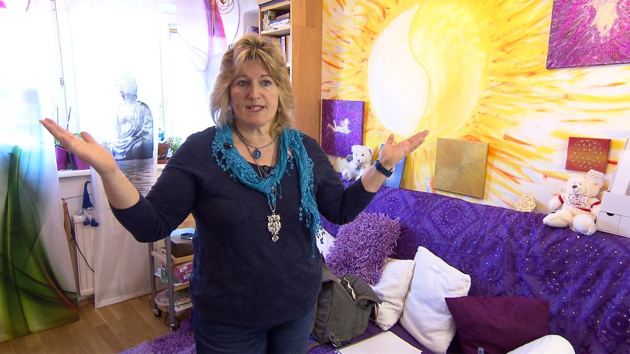 Auf dem Bild ist eine Frau zu sehen, die in einem hellen Raum steht. Sie hat schulterlange, blonde Haare und trägt ein dunkles Oberteil mit einem türkisfarbenen Schal. Ihre Arme sind ausgebreitet, und sie scheint etwas zu erklären oder zu präsentieren.   Im Hintergrund ist eine farbenfrohe Wandgestaltung zu erkennen, die eine strahlende Sonne in Gelb- und Orangetönen zeigt, sowie mehrere gemalte Bilder in verschiedenen Farben. In der Nähe der Wand sind einige kleine Pflanzen in Töpfen sichtbar. Auf einem Sofa im Raum liegen Kissen in Lila, und ein plüschiger Teddybär sitzt darauf. Der Boden ist aus hellem Holz.   Die Atmosphäre wirkt gemütlich und einladend, mit einer spirituellen Note, die durch die Wandgestaltung und die Dekoration des Raumes verstärkt wird.