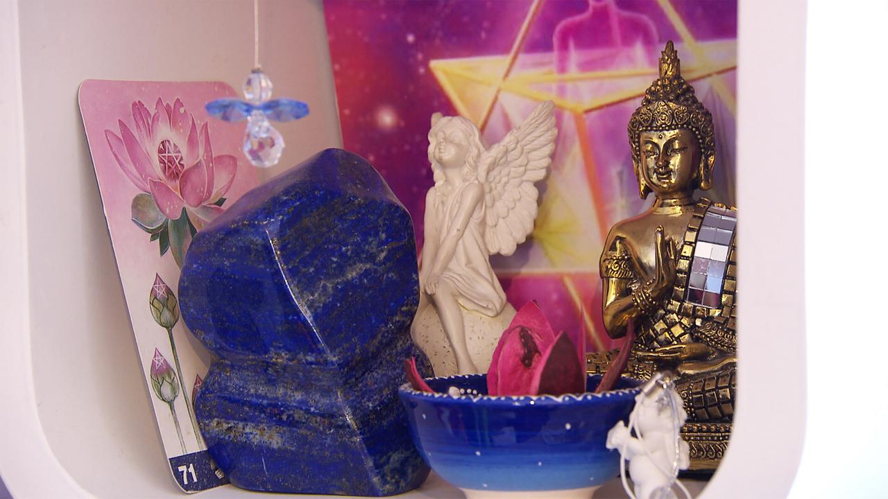 Das Bild zeigt eine dekorative Anordnung, die mit esoterischen und spirituellen Elementen gestaltet ist. Auf der linken Seite befindet sich eine Karte mit einer stilisierten Abbildung einer Lotusblüte in Pink und Grün, die die Nummer 71 trägt. Neben der Karte liegt ein großer, blauer Kristall oder Stein, dessen Oberfläche glänzt und unregelmäßig geformt ist.  In der Mitte des Bildes steht eine kleine, weiße Figur eines Engels, der mit Flügeln versehen ist und eine verträumte Pose einnimmt. Neben dieser Figur befindet sich eine kleine, blaue Schale, die mit getrockneten Blütenblättern gefüllt ist.   Auf der rechten Seite ist eine goldene Buddha-Figur dargestellt, die eine Hand in einer Geste des Segens hebt. Im Hintergrund sind farbige, abstrakte Muster zu sehen, die in warmen Tönen gehalten sind und ein harmonisches Ambiente vermitteln.   Die gesamte Anordnung ist in einem hellen Regal platziert, das dem Arrangement eine ordentliche und zen-artige Ausstrahlung verleiht.