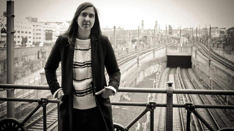 Die Saxophonistin Sarah Chaksad auf einer Bahnhofsbrücke, beide Hände seitlich in den Hosentaschen vom Körper, in die Kamera schauend. Hinter ihr die Gleise.