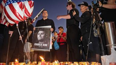 Männer in schwarzer Kleidung und mit US-Flaggen haben sich um Gedenkkerzen und das Schwarz-weiß-Foto eines Mannes versammelt.