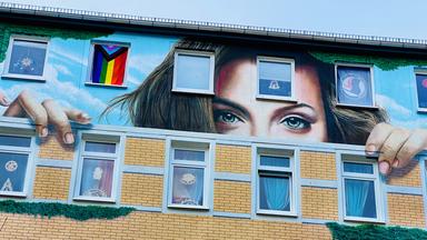 Auf das oberste Stockwerk eines mehrstöckigen Wohnhauses ist die obere Hälfte eines Frauengesichts zu sehen.