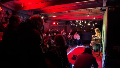 In einem rot ausgeleuchteten Kellerraum spielt eine Person auf der Bühne Gitarre, während rundrum Menschen im Halbdunklen sitzen und zuhören.