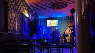 In einem blau ausgeleuchteten Raum stehen drei Personen auf einer Bühne und machen Musik.