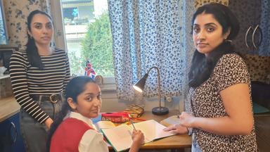 Szene aus "Western Lane" von Chetna Maroo. Ein junges Mädchen sitzt an einem Schreibtisch mit einem aufgeschlagenen Heft und einem Stift in der Hand. Rechts und links von ihr stehen zwei jugendliche Frauen mit einem ernsten Gesichtsausdruck.