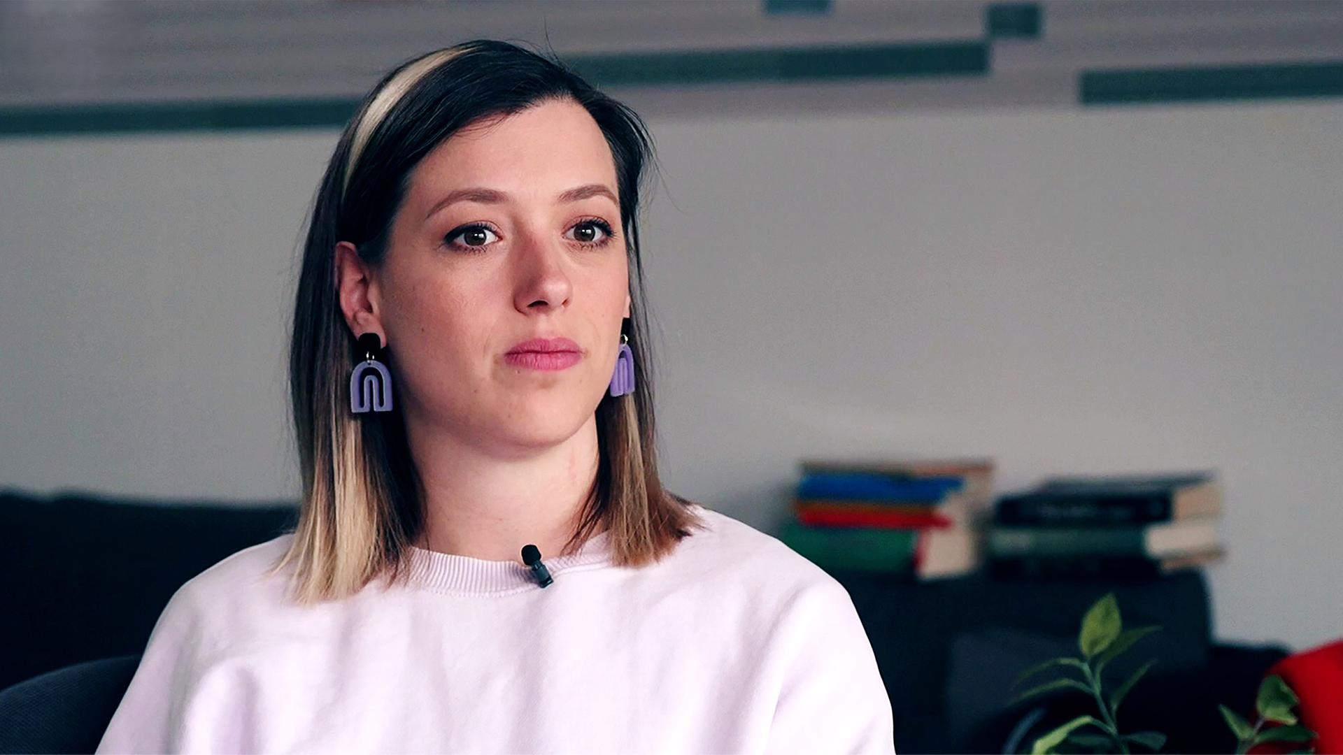 Eine traurig blickende Frau in einem rosafarbenen Pulli und mit lila Ohrringen.