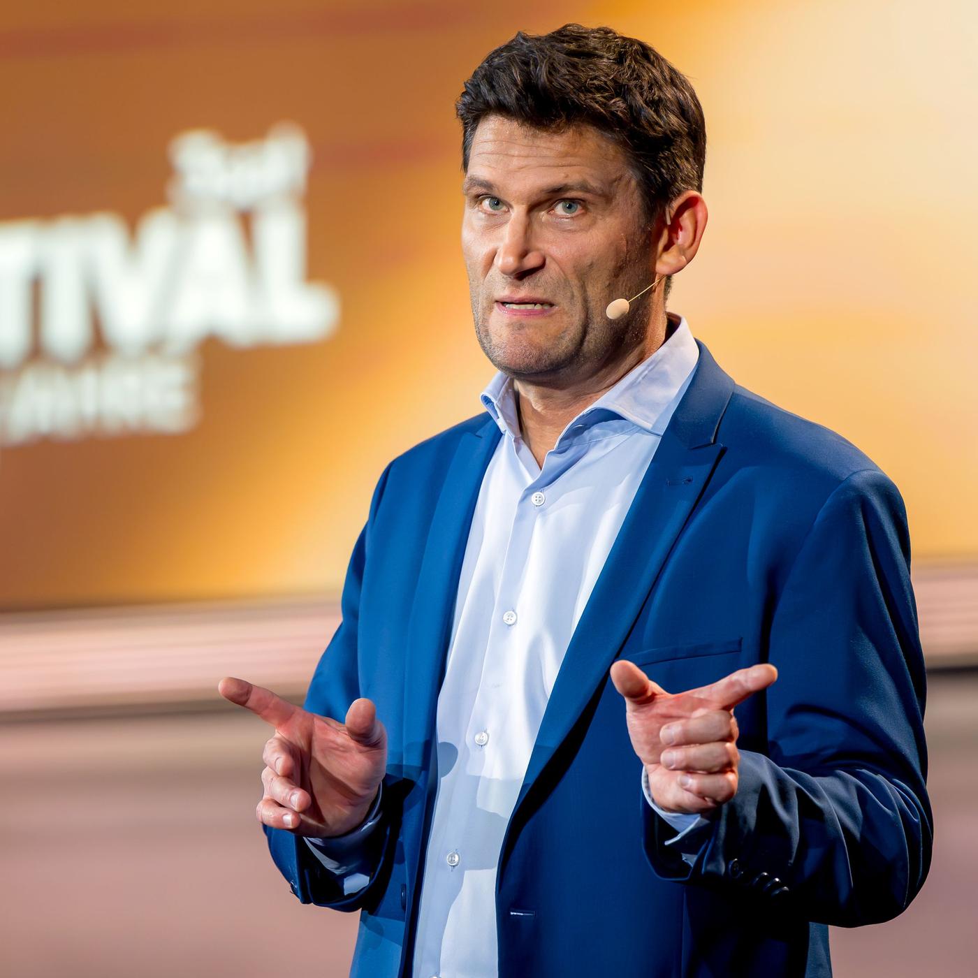 Christian Ehring steht auf der rechten Seite des Bildes. Er hebt beide Zeigefinger. Der Hintergrund ist gelb und braun beleuchtet. Auf der linken Bildseite ist im Hintergrund das 3satFestival Logo abgebildet. Christian Ehring trägt einen dunkelblauen Anzug.