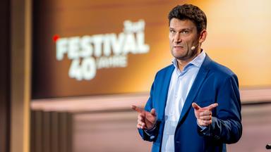 Christian Ehring steht auf der rechten Seite des Bildes. Er hebt beide Zeigefinger. Der Hintergrund ist gelb und braun beleuchtet. Auf der linken Bildseite ist im Hintergrund das 3satFestival Logo abgebildet. Christian Ehring trägt einen dunkelblauen Anzug.