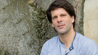 Ein Mann im blauen Shirt und mit braunen Haaren sitzt vor einem Baum und blickt in Richtung des Betrachters.