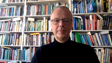 Ein Mann mit schwarzem Pullover und Brille sitzt vor einem Bücherregal.