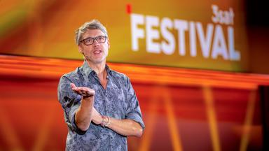 Kabarettist steht auf der linken Seite des Bildes. Er trägt eine Brille. Seine rechte Hand streckt er nach vorne. Er trägt ein gemustertes Hemd. Der Hintergrund ist orange und rot beleuchtet.