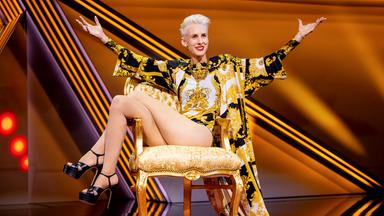 Kabarettistin itzt in der Mitte des Bildes auf einem goldenen Stuhl. Sie breitet die Hände zur Seite aus und streckt die Beine zur linken Bildseite. Der Hintergrund ist orange beleuchtet.