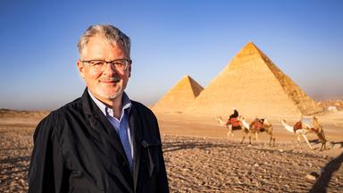 Außenaufnahme von den Pyramiden von Gizeh. Moderator Christopher Clark lächelt in die Kamera. Im Hintergrund ein Mann mit drei Dromedaren.