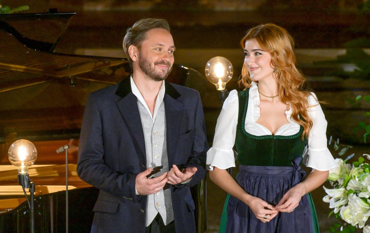 Das Bild zeigt zwei Personen, die auf einer Bühne stehen. Im Hintergrund befindet sich ein Klavier und dekorative Beleuchtung.   Die erste Person, ein Mann, trägt einen dunklen Blazer mit einem hellen Hemd und hat einen Bart. Er hält seine Hände leicht geöffnet vor sich und hat einen freundlichen Gesichtsausdruck.  Die zweite Person, eine Frau, trägt ein traditionelles Outfit, bestehend aus einer dunklen Weste und einem weißen Oberteil mit weiten Ärmeln. Ihr langes, welliges Haar ist braun, und sie lächelt ebenfalls freundlich. Ihre Hände sind vor ihrem Bauch gefaltet.   Um die beiden Personen herum sind einige dekorative Pflanzen und Blumen zu sehen. Die Szene wirkt festlich und einladend, passend zum weihnachtlichen Anlass des Christtagskonzertes.