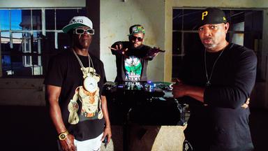 Drei schwarze Männer, die Baseballcaps und teilweise Sonnenbrillen tragen, posieren rund um ein Mischpult.