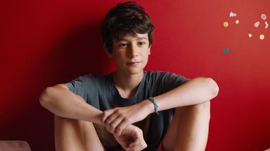 Der 13jährige Rémi sitzt mit angewinkelten Beinen auf dem Bett vor einer rot gestrichenen Wand.