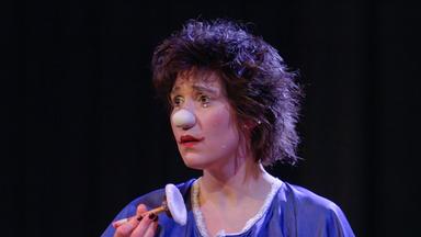 Porträt einer Clownin auf der Bühne im Kostüm mit weisser Clownsnase und Schminke; sie hält einen glitzernden runden Gegenstand in der Hand.