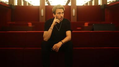 Clueso sitzt auf rotem Sitzpolster und hat die Füße auf dem Tisch vor ihm abgestellt.