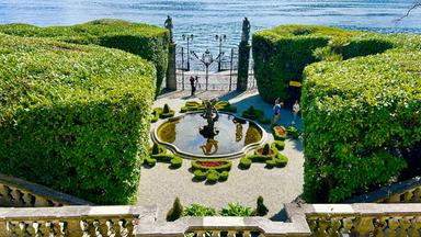 Das Bild zeigt den Garten der Villa Carlotta am Comer See. Im Vordergrund sind Hecken und ein runder Brunnen mit Pflanzen und Figuren zu sehen. Im Hintergrund erstreckt sich der See.