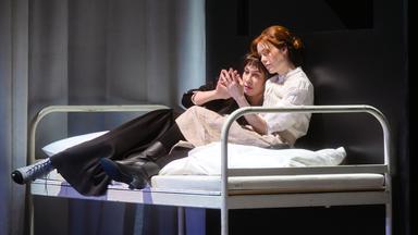 Zwei Frauen liegen in einem Bett auf der Bühen und berühren sich mit einer Hand.