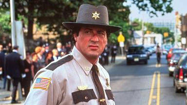 Sheriff Freddy Heflin steht in Dienstkleidung auf einer Straße und blickt zur Seite. Im Hintergrund fahren mehrere Autos, während Menschen am Straßenrand stehen.