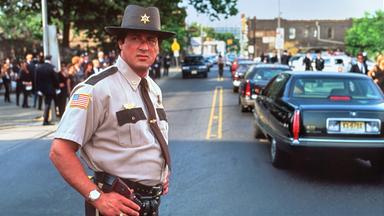 Sheriff Freddy Heflin steht in Dienstkleidung auf einer Straße und blickt zur Seite. Im Hintergrund fahren mehrere Autos, während Menschen am Straßenrand stehen.
