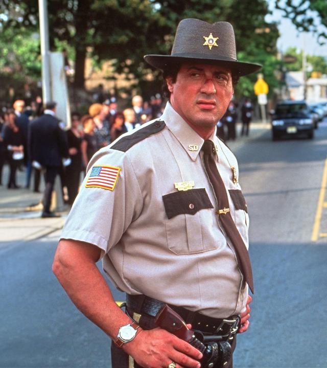 Sheriff Freddy Heflin steht in Dienstkleidung auf einer Straße und blickt zur Seite. Im Hintergrund fahren mehrere Autos, während Menschen am Straßenrand stehen.