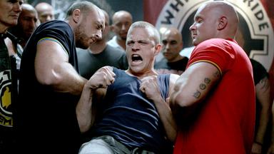 Das Bild zeigt eine Szene aus dem Film "CopStories – Bist du deppert". In einem dunklen und angespannten Raum stehen mehrere muskulöse Männer, die intensiv in eine Auseinandersetzung verwickelt sind.   Im Vordergrund sieht man einen jungen Mann mit kurzgeschorenen Haaren, der ein ärmelloses Oberteil trägt. Sein Gesicht ist verzerrt vor Wut oder Anstrengung, während er mit beiden Fäusten in der Luft kämpferisch wirkt. Er wird von zwei kräftigen Männern festgehalten, einer von ihnen trägt ein dunkelblaues Shirt mit gelben Akzenten, der andere ein rotes Shirt und hat sichtbare Tätowierungen auf seinem rechten Arm.  Im Hintergrund sind weitere Männer zu sehen, die aufmerksam auf die Szene reagieren; sie sind nahezu alle glatzköpfig oder haben sehr kurze Haare. Der Raum hat eine düstere Atmosphäre, mit wenig Licht und einer Wand, die mit dem Schriftzug "WHITE PRIDE" dekoriert ist.   Die gesamte Komposition des Bildes vermittelt ein Gefühl von Aggression und Konflikt, und die Körperhaltungen der Figuren zeigen, dass sich kurz vor einem entscheidenden Moment etwas anbahnt.