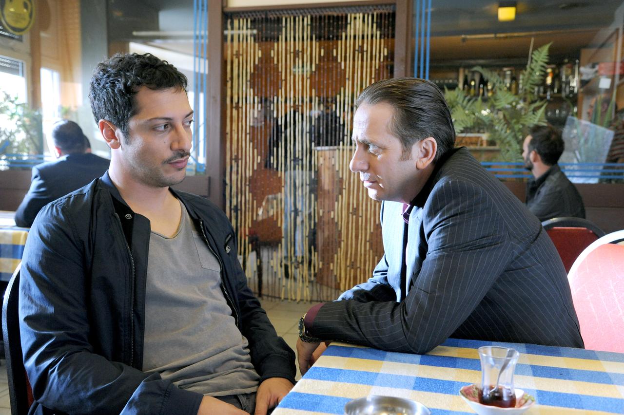 Das Bild zeigt zwei Männer, die sich in einem Restaurant oder einem Café unterhalten. Der Mann links hat kurze, dunkelbraune Haare und trägt eine schwarze Jacke über einem grauen T-Shirt. Er sieht nachdenklich aus und hat einen leichten Schnurrbart. Der Mann rechts hat ebenfalls kurze Haare, ist ordentlich gekleidet in einem gestreiften Anzug und einem lila Hemd. Er beugt sich leicht über den Tisch, während er intensiv mit dem anderen spricht.   Im Hintergrund sind weitere Personen zu sehen, die ebenfalls in dem Lokal sitzen. Die Tischdecke ist blau mit gelb karierten Mustern. Auf dem Tisch stehen ein Glas Tee und ein Teller mit kleinem Gebäck. An den Wänden sind dekorative Elemente, einschließlich Pflanzen und einer Art Wandverkleidung zu sehen, die dem Raum eine gemütliche Atmosphäre verleiht.