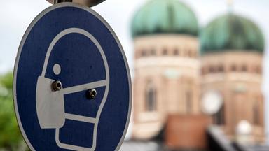 Auf einem runden Verkehrsschild ist ein schematisches Bild eines Kopfes mit Mund-Nase-Bedeckung zu sehen.